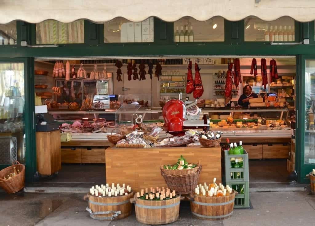 Wine bottles in baskets on the ground and assorted meats hang from hooks at this outdoor food stand. 