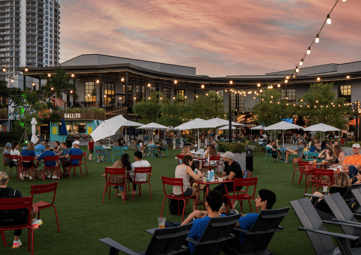 People sitting in lawn chairs at sunset eating and drinking with strand lights above them.